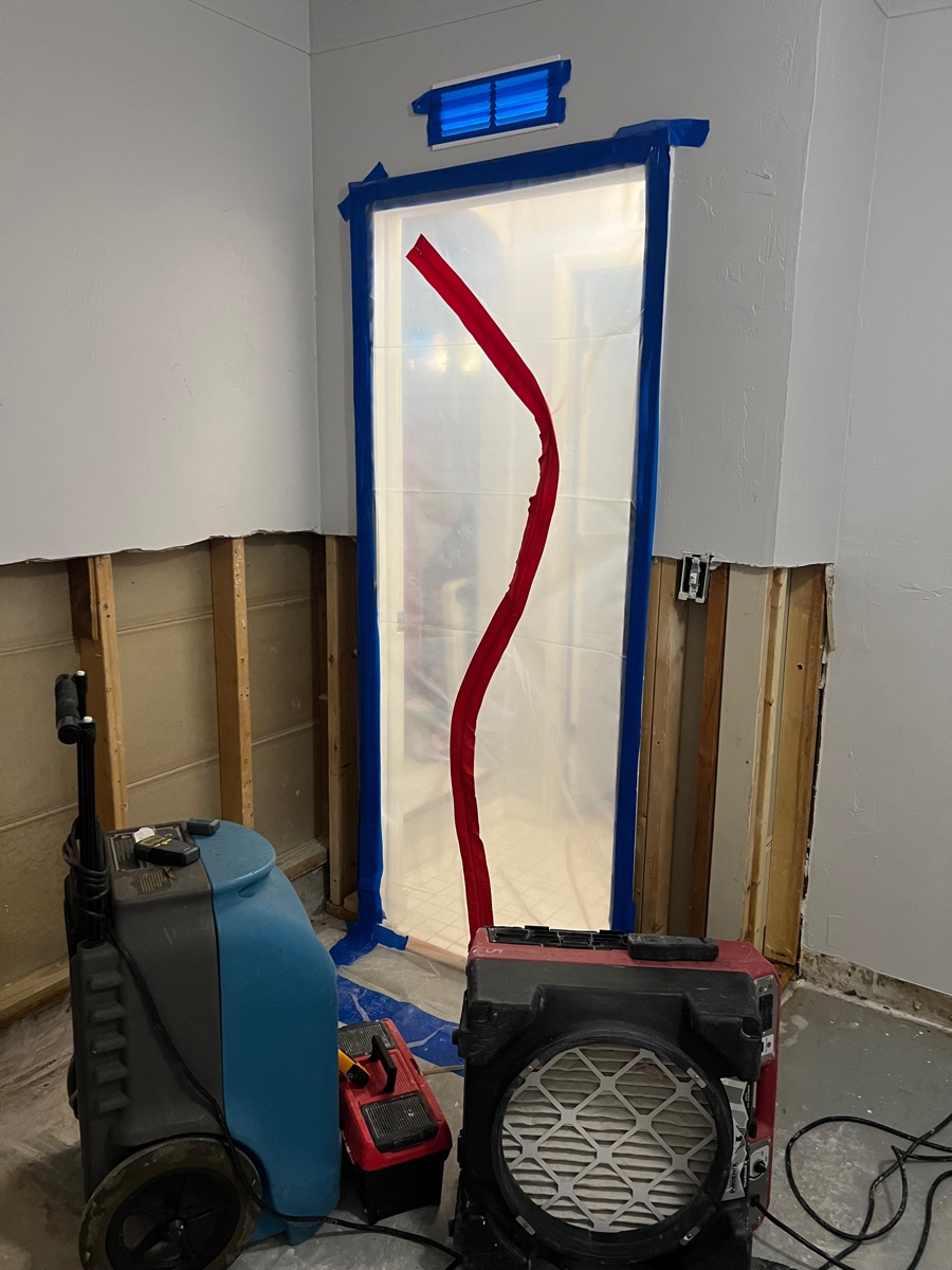 Plastic sheeting containment with a zipper door, with a dehumidifier and air mover in front of walls with the lower drywall removed